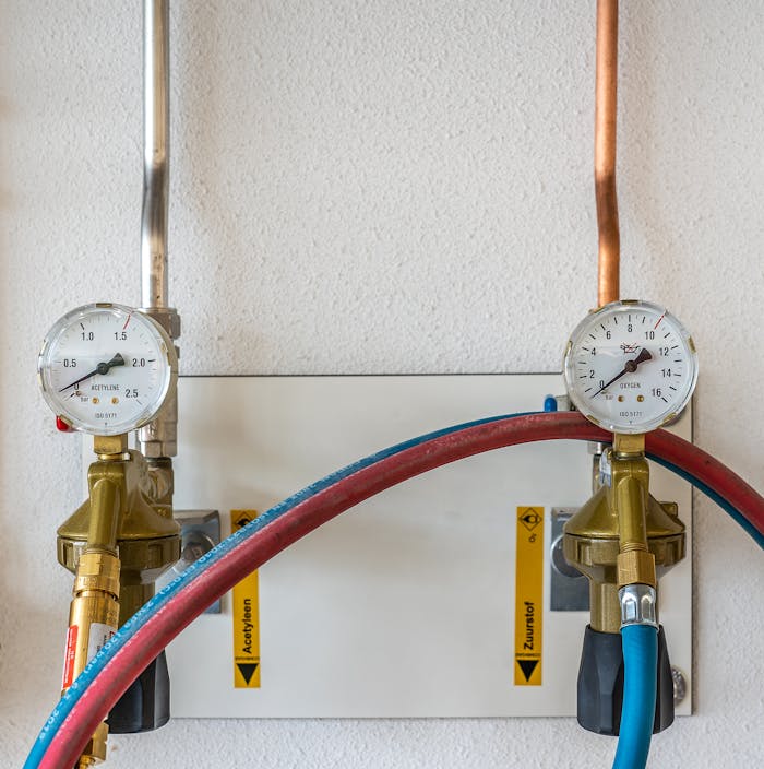 Close-up of industrial gas pressure gauges connected with hoses against a white wall.